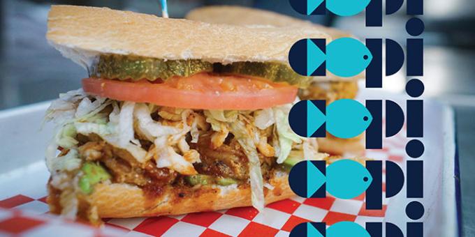 A tall layered sandwich with pickles, tomato, shredded lettuce and fish with a dark sauce.