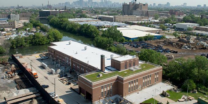A red brick building with grass on the roof sits on the edge of a small river. A city skyline is seen in the distance.
