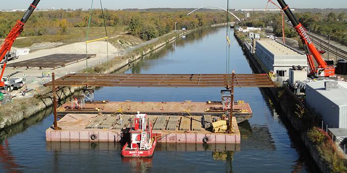 A two cranes standing on shore move electrical barrier equipment from a barge into water.