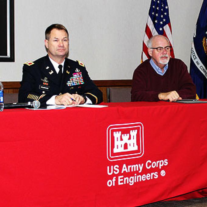 Three men sit at a long table draped with a cloth displaying the U.S. Army Corps of Engineers' logo.