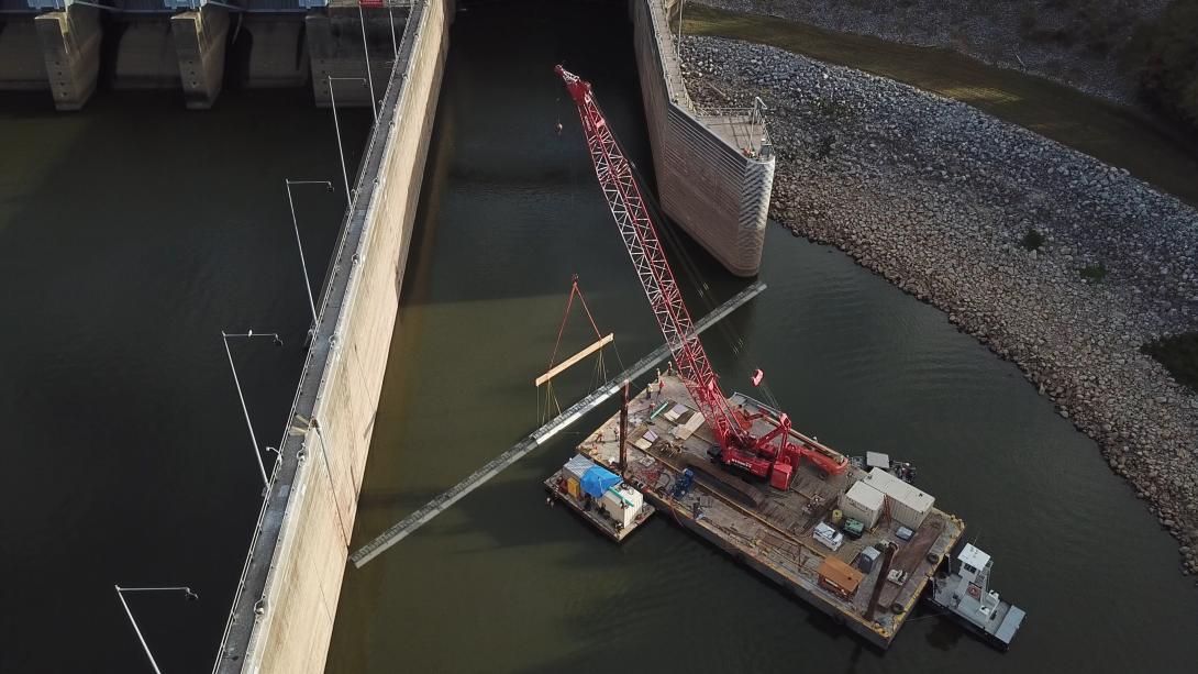 A huge red crane on a platform in the water installing a section of fish fence at a dam.