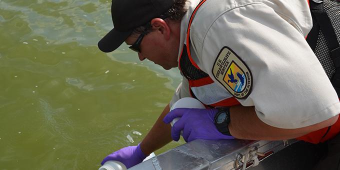 A biologist wearing latex gloves collects water in a plastic container over the side of a boat.