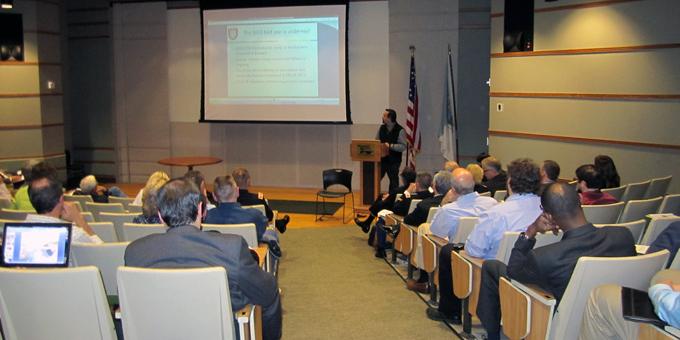 People sitting in an auditorium looking toward a presenter and projection screen.
