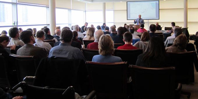 A group of people sits in a large conference room looking forward at a presenter standing in front of a projector screen.