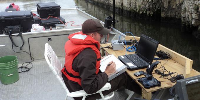 A person holding a clipboard with papers sits on a boat looking at a laptop computer sitting on a makeshift desk.