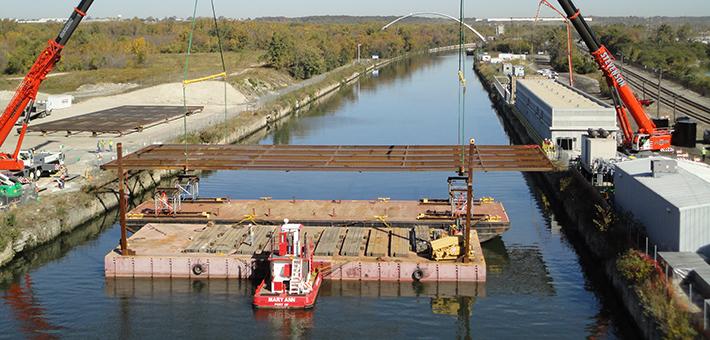 A two cranes standing on shore move electrical barrier equipment from a barge into water.